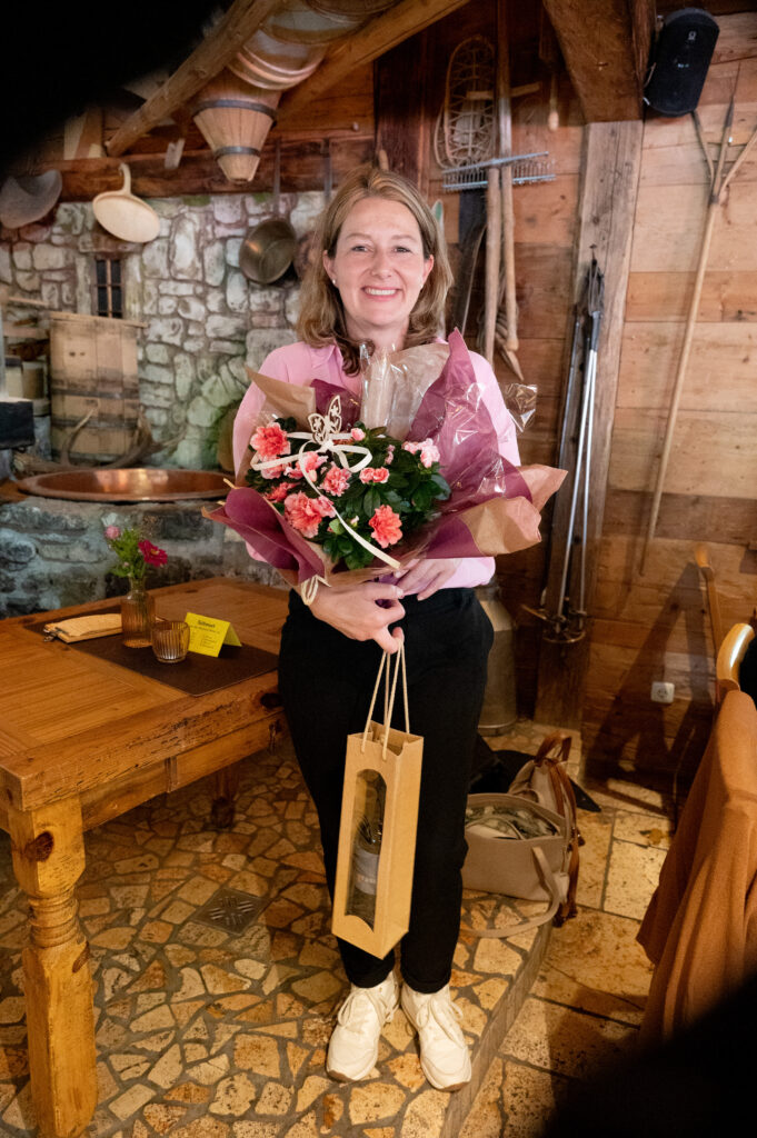 Violetta Giselbrecht hält einen Blumenstrauß und eine Geschenksttasche mit einer Flasche drinnen in der Hand.