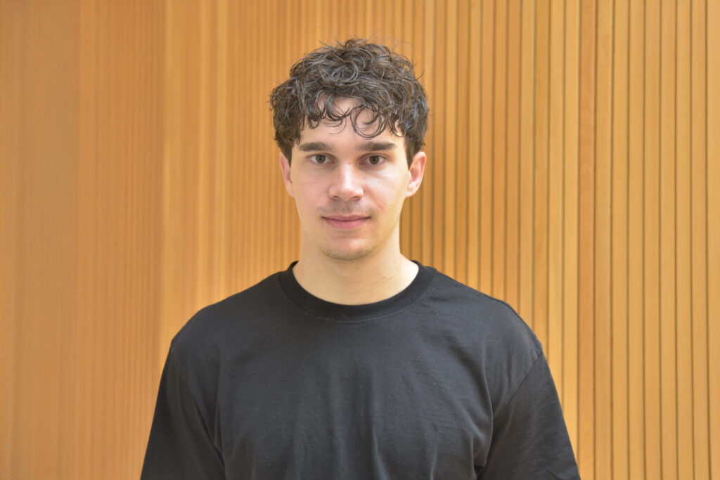 Lukas Pienz steht vor einer Holzwand, hat lockige Haare und ein trägt ein schwarzes T-Shirt.