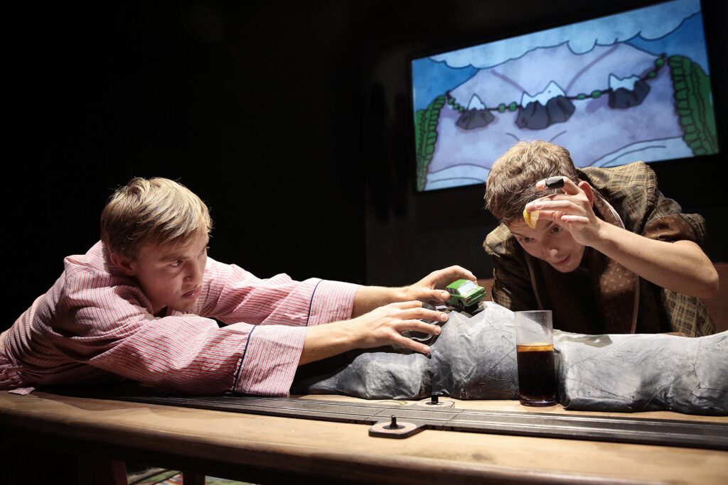 Ein Schauspieler fährt mit einem Spielzeugauto über den Tisch und ein anderer träufelt Zitrone in ein Glas Cola.