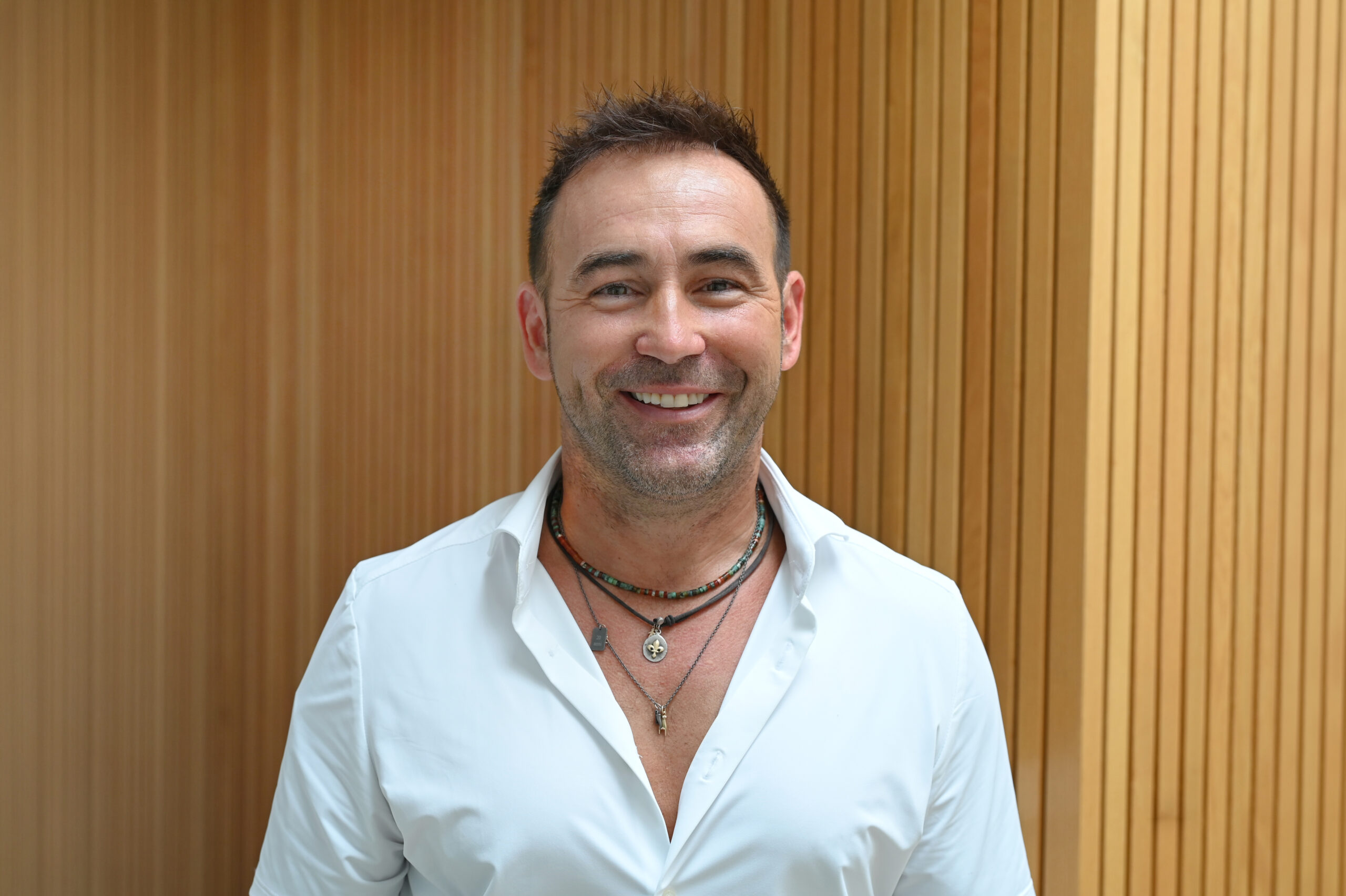 Ein Portrait von Thomas König, der lacht und ein weißes Hemd und Ketten trägt.