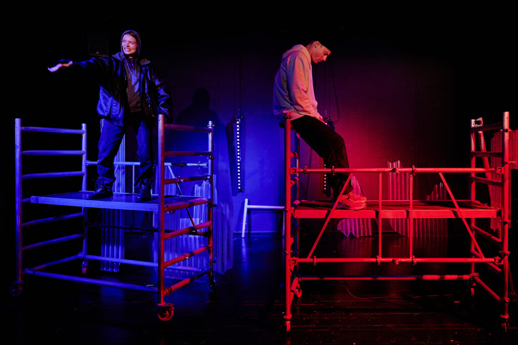 Das Bild zeigt eine Schauspielerin und und ein Schauspieler auf Gerüsten mit blauer und roter Beleuchtung.